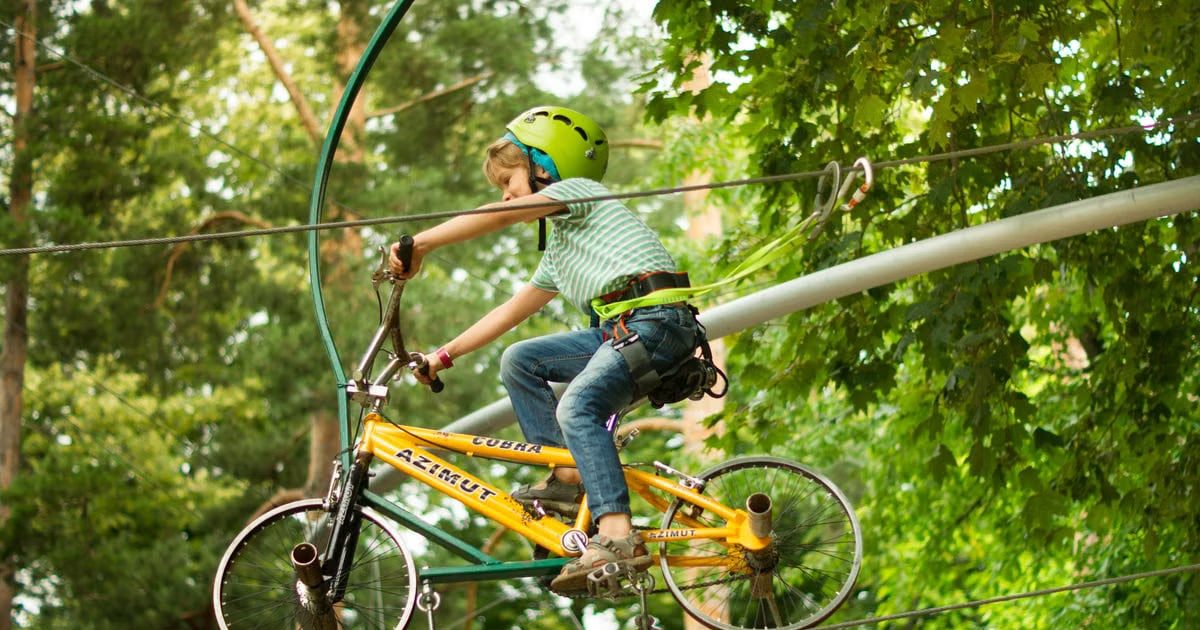 A child wearing a helmet rides a bike on a zipline in an adventure park surrounded by trees.