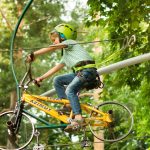 A child wearing a helmet rides a bike on a zipline in an adventure park surrounded by trees.