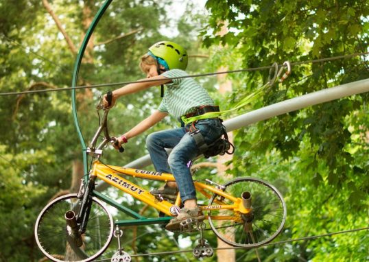A child wearing a helmet rides a bike on a zipline in an adventure park surrounded by trees.