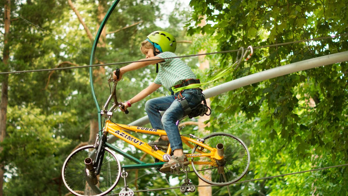 A child wearing a helmet rides a bike on a zipline in an adventure park surrounded by trees.